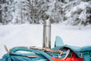 A bright hiking backpack lies on the snow. A climbing rope is tied to a backpack. In the side pocket of the thermos. Hiking in winter. Rocks and forests photo