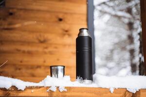 black matte thermos and metal mug with hot drink on window sill of the outdoor veranda. Hiking and picnic staff. Drinking warm tea or coffee in the winter frost. photo