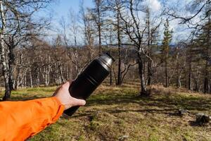 A person holds a black thermos in his hand against the background of the forest, a tourist shows a vacuum bottle for tea, a thermos with a hot drink, equipment for hiking in nature. photo