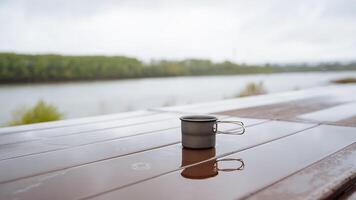 café jarra en naturaleza, cámping utensilios, Bebiendo caliente té en un caminata, reflejando el silueta de un vaso en un charco de agua, un taza en pie en el mesa húmedo, lluvia en el bosque foto