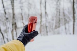 A gloved hand holds a red thermos. Thermo mug in hand. Close-up shots against the background of the forest. photo