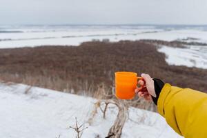 un naranja jarra en tu mano, té vertido dentro un taza, sostener un vaso lleno con un caliente beber, un invierno caminata en el bosque, un paisaje de hermosa naturaleza. foto