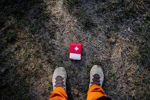 First aid kit lies on the ground at the feet, top view red bag of medicines, camping equipment, hiking, survival in the forest, cross white, photo