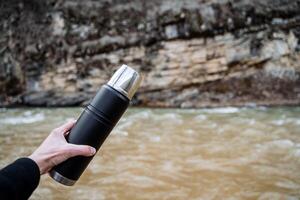 un vacío termo sostiene su mano en contra el antecedentes de el río. el concepto de cámping utensilios para un turista, un botella para caliente bebidas, un termo con té. foto