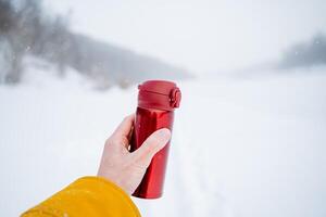 A person holds a thermos with a hot drink in his hand. The concept of winter recreation in nature. photo