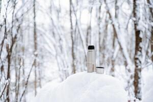 An iron thermos of silver color stands on the snow. Thermo mug for hot drinks. Winter in the forest, hot coffee in a glass. photo