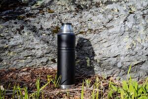 Black thermos stands on the background of a stone, vacuum bottle for coffee, a vessel for a hot drink, no lid, green grass, a piece of rock, metal steel photo