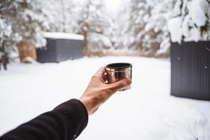 un metal taza con té en el manos de un viajero. bebida calentar té y café desde un termo en un caminar en invierno. cubierto de nieve bosque en un tranquilo día. brillante ropa foto