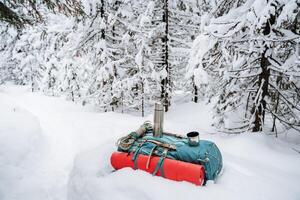 View of a backpack lying in the snow near the trail. The red mat is fastened with straps to the backpack. Thermos of silver color. A harsh winter hike through the forest. photo