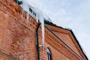 A huge icicle hangs from the roof of a brick building. Dangerous and sharp icicles on the facade of the house. Be careful in winter.natural phenomena. photo