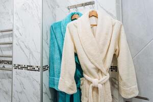 A terry robe hangs on a hanger in the bathroom, two warm bathrobes after a shower, clothes absorb moisture from the body, a belt on a bathrobe, a set of clothes in a hotel. photo