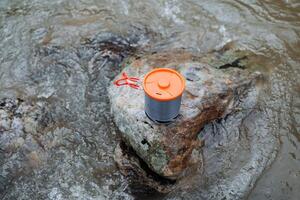 Tourist tableware concept on a stone against the background of water, a pot stands on a stone in the river, an orange cup lid, folding handles. photo
