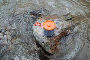 turista vajilla concepto en un Roca en contra el antecedentes de agua, un maceta soportes en un Roca en el río, un naranja taza tapa, plegable manejas. foto