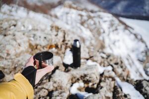 Thermos with hot tea to drink in the mountains in nature. Winter hike of a tourist on the rocks. Drink coffee in the morning in nature. Hiking. photo