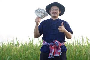 Asian man farmer is at paddy field, holds Thai banknote money and thumb up. Concept , Happy farmer get profit, income, agriculture supporting money. Proud on crops. photo