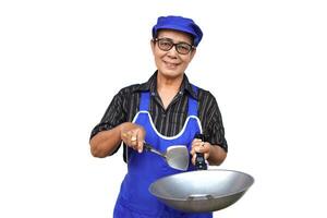 Asian senior woman, wears blue apron and cap, holds frying pan and spatula or ladle. Look at camera, smiles, feels confident isolated on white background. Concept, love cooking. Kitchen lifestyle. photo