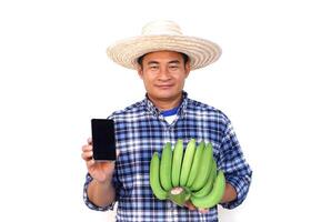 hermoso asiático hombre agricultor, usa sombrero, azul tartán camisa, sostiene teléfono inteligente y verde plátanos, aislado en blanco antecedentes. concepto, agricultura ocupación. tailandés granjero crece orgánico bananas para de venta foto