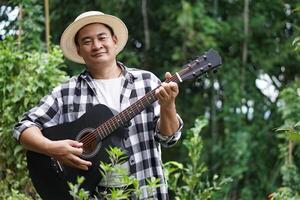 Handsome Asian man is playing acoustic guitar in the park, outdoor nature background. Concept, love music, hobby, recreation activity. Relax time. Music can develop mental health. photo