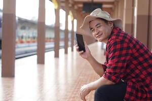 Asian man traveler is waiting someone at platform of train station, holds mobilephone to talk on the phone. Concept, travel alone, transportation, communication or connection photo