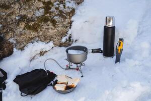 Lunch in nature, cooking in camping conditions, winter camping, sandwiches lying on a plate, melting snow in a pot on a burner. thermos with tea. photo