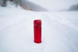 A red thermos on the background of snow. Winter photo of thermo mugs. A glass for hot drinks.