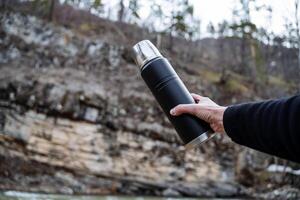 A tourist person holds a vacuum thermos in his hand against the background of a rock. Black thermal bottle for hot drinks. photo