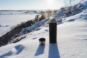 The concept of a thermos against the background of winter nature. A black thermos with a mug stands in the snow. Sunlight, shade from the object, tourist equipment. photo