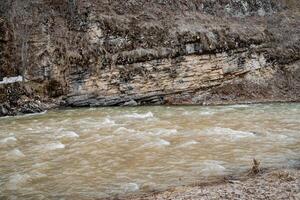 Spring flood on a mountain stream, the river flows at the foot of the rock, brown color of the water in spring, cloudy day. photo