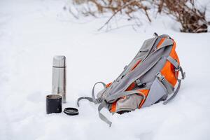 Thermos with hot tea. The backpack is lying in the snow. Tourist things stand in the snow. Metal tableware for camping. photo
