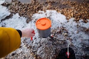 Hand holds the handle of the boiler on the gas burner on the hike, the tourist cooks food on the burner, winter hike ice on the ground, tourist dishes, light metal alloy photo