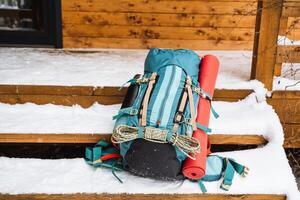 A bright hiking backpack lying on the porch near the house. A climbing rope is tied to a backpack. In the side pocket of the thermos. Hiking in winter. Rocks and forests photo