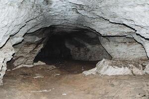 paso subterráneo en un Roca cueva, un karst cavidad en el ladera de la montaña, oscuridad a el final de el túnel, arcilla piso, gris caliza paredes foto