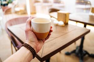 A hand holds a mug of tea in front of the cafe. Drink a hot drink at the table in the restaurant. Glass in hand bracelet, thumb. Aroma of masala tea. photo