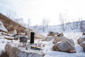 camping utensils standing on a stone, camping equipment, gas burner for hiking, thermos, kettle with tea, pan cooking, cold winter forest stones photo