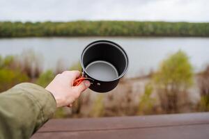 un persona sostiene un vacío maceta en su mano, un maceta para comiendo en un caminata es Disparo de cerca, cámping utensilios, un bosque río, un viaje, trekking en naturaleza. foto