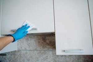 Cleaning of cabinets in the kitchen with a damp cloth. Wipe door handles with detergent. Disinfection of surfaces. Hand with rag. Blue glove. photo