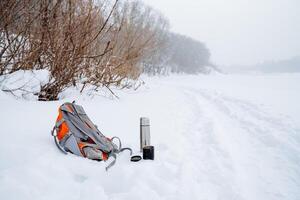 The backpack lies in the snow near the road. The winter trail runs along the river. Equipment for a winter hike. thermos with hot tea. photo