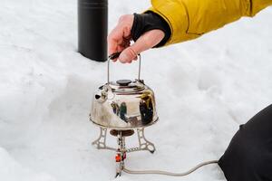 A tourist in a hike puts a kettle of water on a gas burner, boil water on fire, brew hot tea, take a pot with his hand, a concept of tourist dishes, a hike photo