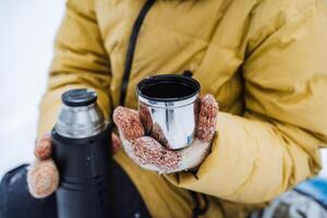 Thermo mug in hand. A hand holds a mug of hot tea. Drink tea in the winter outside. A metal cup with a drink is held by a person. Tea from a thermos. photo