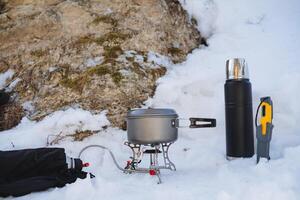 Tourist utensils, cooking in nature in the winter season, a burner with a pan stands on the snow, a thermos with tea, a bushcraft knife. photo