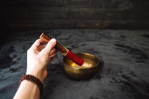 un mano sostiene un gong palo, un canto cuenco soportes lado por lado en el cama. un objeto ese hace el sonidos de un campana, meditación en silencio. dorado metal musical instrumento. foto