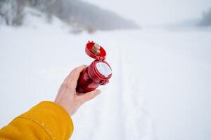 Red thermos with lid open. Hold a thermos for tea in your hand. Close-up shots against the background of snow. photo