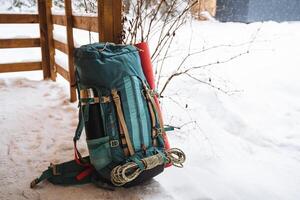 A bright hiking backpack lying on the porch near the house A climbing rope is tied to a backpack. In the side pocket of the thermos and mat for sleeping. Hiking in winter. Rocks and forests photo