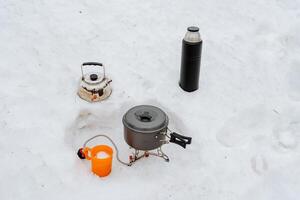 Tourist utensils set stands on the snow, the concept of cooking in nature in a campsite in winter, a thermos with hot tea, a pot of porridge, a metal kettle for water. photo