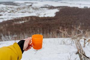 A person holds a glass in his hand, drink tea against the background of the forest, an orange cup is filled with tea, outdoor recreation, dishes in his hands photo