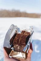 Chocolate against the background of white snow. Slices of milk chocolate are held in the hands of a person. A sweet snack in the woods. Campaign. photo