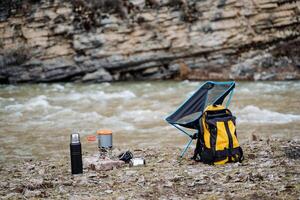turista acampar en el bancos de un montaña río, amarillo mochila soportes en el suelo, plegable ultraligero silla, cámping utensilios, almuerzo en naturaleza, termo con té, maceta foto