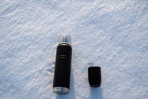 A new black thermos and a glass lie in the snow. Vacuum cup for a hot drink. Tea in a thermos in winter. photo