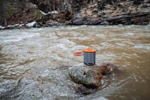 Tourist utensils stand on a stone in the river, spring hike in the mountains, a pot with a lid and folding handles, light equipment, compact size of dishes. photo