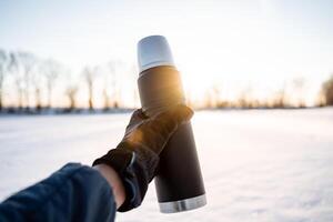 Steel vacuum thermos hold in your hand in nature. Black thermos with metal cup. Hot tea in nature in the rays of the setting sun. photo